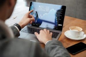 Ein Investor in Business-Kleidung analysiert auf einem Laptop eine Börsenkursgrafik mit steigenden und fallenden Kurven. Er zeigt mit dem Finger auf einen bestimmten Punkt des Charts. Auf dem Holztisch stehen eine Tasse Cappuccino und ein Smartphone, was eine moderne Arbeitsatmosphäre vermittelt.