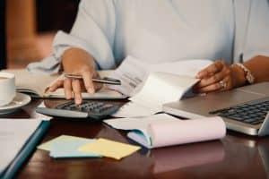 Eine Person sitzt an einem Schreibtisch und bearbeitet finanzielle Unterlagen. Sie verwendet einen Taschenrechner, hält einen Stift in der Hand und blättert durch Quittungen und Dokumente. Auf dem Schreibtisch liegen ein Laptop, bunte Haftnotizen, ein Notizbuch und eine Tasse Kaffee.