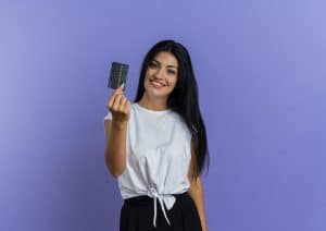 Eine lächelnde Frau mit langen dunklen Haaren hält eine Kreditkarte in der Hand und präsentiert sie stolz vor einem lila Hintergrund. Sie trägt ein weißes T-Shirt und schwarze Hosen, was einen lässigen, aber selbstbewussten Eindruck vermittelt. Die Szene symbolisiert finanzielle Freiheit und den bewussten Umgang mit Kreditkarten im Alltag.