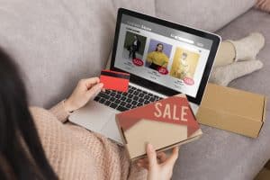 Eine Frau sitzt bequem auf einem Sofa, hält eine Kreditkarte in der Hand und tätigt Online-Käufe über ihren Laptop. Auf dem Bildschirm ist ein Modegeschäft mit Rabattangeboten zu sehen, während sie gleichzeitig ein Paket mit der Aufschrift "SALE" hält. Die Szene symbolisiert den Komfort und die Beliebtheit des Online-Shoppings in der heutigen digitalen Welt.