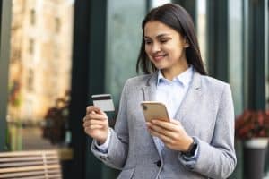 Eine junge Frau in einem grauen Blazer steht lächelnd im Freien vor einem Gebäude, hält in der einen Hand eine Kreditkarte und in der anderen ein Smartphone, während sie offenbar eine Online-Zahlung oder einen digitalen Kauf tätigt.