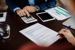 Zwei Personen arbeiten an einem Schreibtisch mit Dokumenten, Taschenrechner, Tablet und Laptop. Eine Person tippt auf dem Taschenrechner, während die andere etwas auf ein Formular schreibt – eine typische Szene, die die Bürokratie im Bankwesen veranschaulicht.