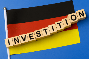 Holzwürfel mit dem Schriftzug „INVESTITION“ liegen auf einer deutschen Flagge vor blauem Hintergrund. Die Szene symbolisiert Kapitalanlagen in Deutschland im aktuellen wirtschaftlichen Umfeld. Besonders relevant ist der Einfluss des EZB-Zinssatzes auf Investitionsentscheidungen und die Finanzmärkte der Eurozone.