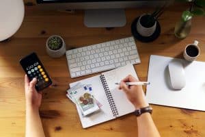 Eine Person sitzt an einem Schreibtisch und beschäftigt sich mit der Finanzplanung. Auf dem Holztisch liegen ein Smartphone mit geöffnetem Taschenrechner, Euro-Banknoten und -Münzen, ein Notizbuch mit Berechnungen, eine Tastatur, eine Maus sowie eine Tasse Kaffee. Die Szene vermittelt den Eindruck einer sorgfältigen Finanzplanung im Homeoffice.
