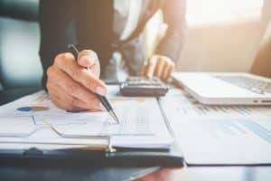 Ein Geschäftsmann arbeitet an der Finanzplanung, während er Finanzdokumente mit Diagrammen und Tabellen analysiert. Er macht Notizen mit einem Stift und benutzt gleichzeitig einen Taschenrechner. Auf dem Tisch liegen ein Laptop und weitere Unterlagen.