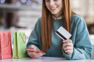 Eine junge Frau sitzt an einem Tisch mit Einkaufstüten, hält eine Kreditkarte in der einen Hand und ihr Smartphone in der anderen. Sie lächelt und scheint einen Online-Kauf zu tätigen. Die Szene symbolisiert modernes Shopping mit Fokus auf digitale Zahlungen und Cashback-Vorteile bei Einkäufen.