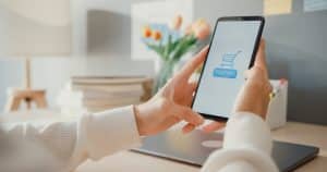 Eine Person hält ein Smartphone mit geöffneter Zahlungsanwendung in der Hand, auf dem ein Einkaufswagensymbol und der Schriftzug „Payment“ zu sehen sind. Im Hintergrund befinden sich ein Schreibtisch, Bücher und ein Laptop. Die Szene zeigt den Einsatz von Karten virtuell für sicheres und bequemes Online-Shopping im häuslichen Umfeld.