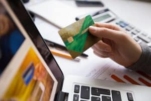 Eine Person hält eine grüne Kreditkarte in der Hand und schaut auf einen Laptop-Bildschirm mit einer E-Commerce-Website. Im Hintergrund liegen ein Taschenrechner, ein Stift und ein Dokument mit einem Balkendiagramm. Das Bild symbolisiert Online-Shopping und digitale Bezahlung mit Kreditkarten.