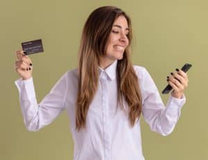 Eine junge Frau mit langen braunen Haaren lächelt, während sie auf ihr Smartphone schaut und in der anderen Hand eine Kreditkarte hält. Sie trägt ein weißes Hemd und steht vor einem olivgrünen Hintergrund. Die Szene vermittelt Freude und Zufriedenheit beim Online-Shopping oder bei der Nutzung von Kreditkartenprogrammen (Kreditkartenprogramme).