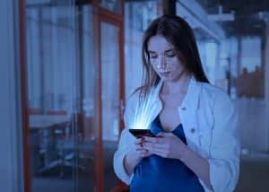 Eine Frau steht in einem modernen Bürogebäude und schaut auf ihr Smartphone, während ihr Gesicht von einem digitalen Raster erfasst wird. Leuchtende Linien symbolisieren die Datenübertragung vom Gerät. Die Szene zeigt den Einsatz von Künstliche Intelligenz zur Gesichtserkennung und biometrischen Authentifizierung im digitalen Alltag.