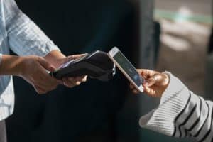 Eine Person hält ein Smartphone über ein Kartenlesegerät, um eine kontaktlose Zahlung durchzuführen. Auf dem Bildschirm des Handys ist ein Symbol für NFC-Zahlung mit der Schaltfläche „PAY“ sichtbar. Die Szene symbolisiert moderne digitale Zahlungen im Einzelhandel. Diese Darstellung steht im Einklang mit den Zielen der neuen PSD3-Richtlinie, die sichere und nahtlose elektronische Zahlungen im europäischen Zahlungsverkehr fördern soll.