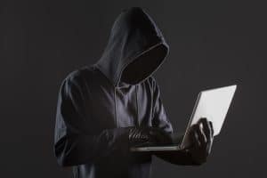 Eine vermummte Person trägt einen dunklen Kapuzenpullover und schwarze Handschuhe, während sie einen Laptop benutzt. Das Gesicht bleibt im Schatten verborgen, was Anonymität suggeriert. Diese Szene symbolisiert typisches Verhalten im Zusammenhang mit digitaler Betrug und verdeutlicht die Bedrohung durch Cyberkriminalität in der modernen digitalen Welt.