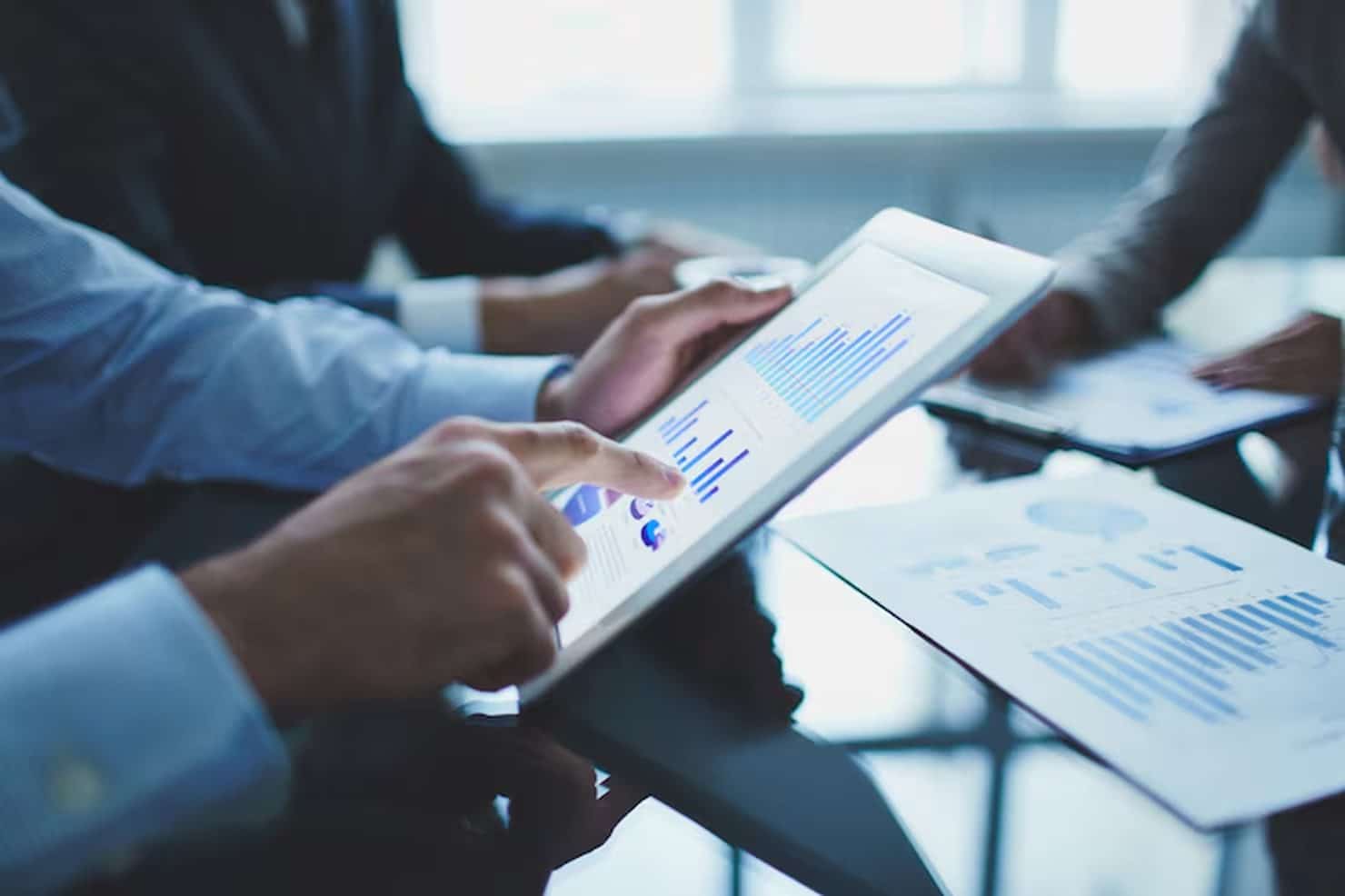 Ein Investor analysiert mit einem Tablet detaillierte Finanzdiagramme, während weitere Investoren im Hintergrund an einem Konferenztisch Daten auf Papier betrachten – ein moderner Geschäftskontext, der den datenbasierten Entscheidungsprozess von Investoren veranschaulicht.