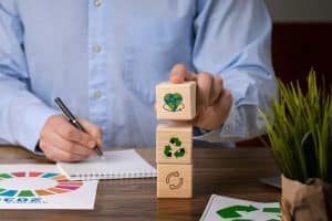 Ein Geschäftsmann im blauen Hemd sitzt an einem Schreibtisch und hält einen Holzbaustein mit einem grünen Herzsymbol in der Hand. Darunter sind weitere Würfel mit Recycling- und Kreislaufsymbolen gestapelt. Auf dem Tisch liegen ESG-Dokumente, ein Notizbuch und eine grüne Pflanze – eine Szene, die das Engagement für nachhaltige Investitionen und umweltbewusstes Wirtschaften visuell darstellt.