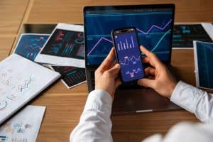 Eine Person analysiert Finanzdiagramme auf einem Smartphone und Laptop, umgeben von gedruckten Berichten mit bunten Linien- und Balkendiagrammen. Die Szene zeigt eine moderne Arbeitsumgebung, in der datenbasierte Entscheidungen für Investitionen getroffen werden – typisch für digitale Finanztrends wie Crowdinvesting.