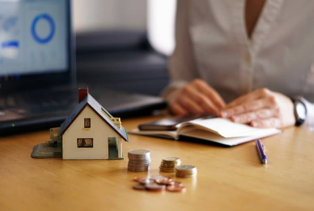 Miniaturhaus, Münzstapel und Notizbuch auf einem Schreibtisch mit Laptop im Hintergrund – Symbol für Finanzplanung und Investitionen in Immobilien.