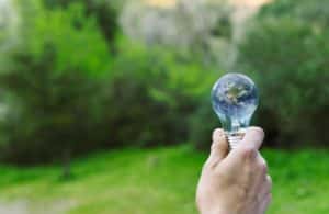Eine Hand hält eine Glühbirne, in der sich die Erde spiegelt, vor einem grünen Hintergrund. Das Bild symbolisiert Saubere Energie, Nachhaltigkeit und den Schutz unseres Planeten.