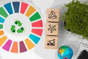Eine symbolische Darstellung der Nachhaltigkeit mit farbigen Segmenten, Holzblöcken mit Umwelt- und Recycling-Symbolen, einer kleinen Weltkugel und einer Pflanze. Im Mittelpunkt stehen Themen wie Kreislaufwirtschaft, Umweltschutz und wirtschaftliches Wachstum im Kontext von Nachhaltigkeitsindizes.
